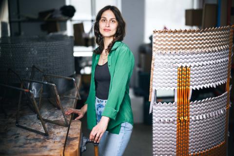 Alina Tenser stands between a rustic wooden workbench holding metal rebar structures and a large, curved sculpture featuring woven metallic and orange ribbons.