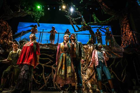 A dramatic stage production featuring a group of diverse actors looking upward with expressions of awe and anticipation. The set design is highly detailed, featuring massive, twisted artificial trees with textured bark, a chain-link fence on a raised platform, and a blue-sky backdrop. Bright green and white spotlights illuminate the performers from above.