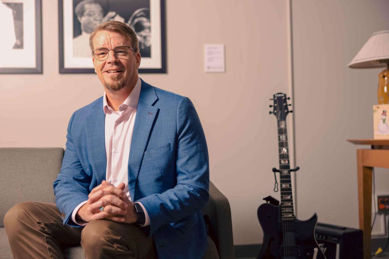 Dean Bob Flowers, sitting on a couch next to his guitar in his office smiling for his portrait.