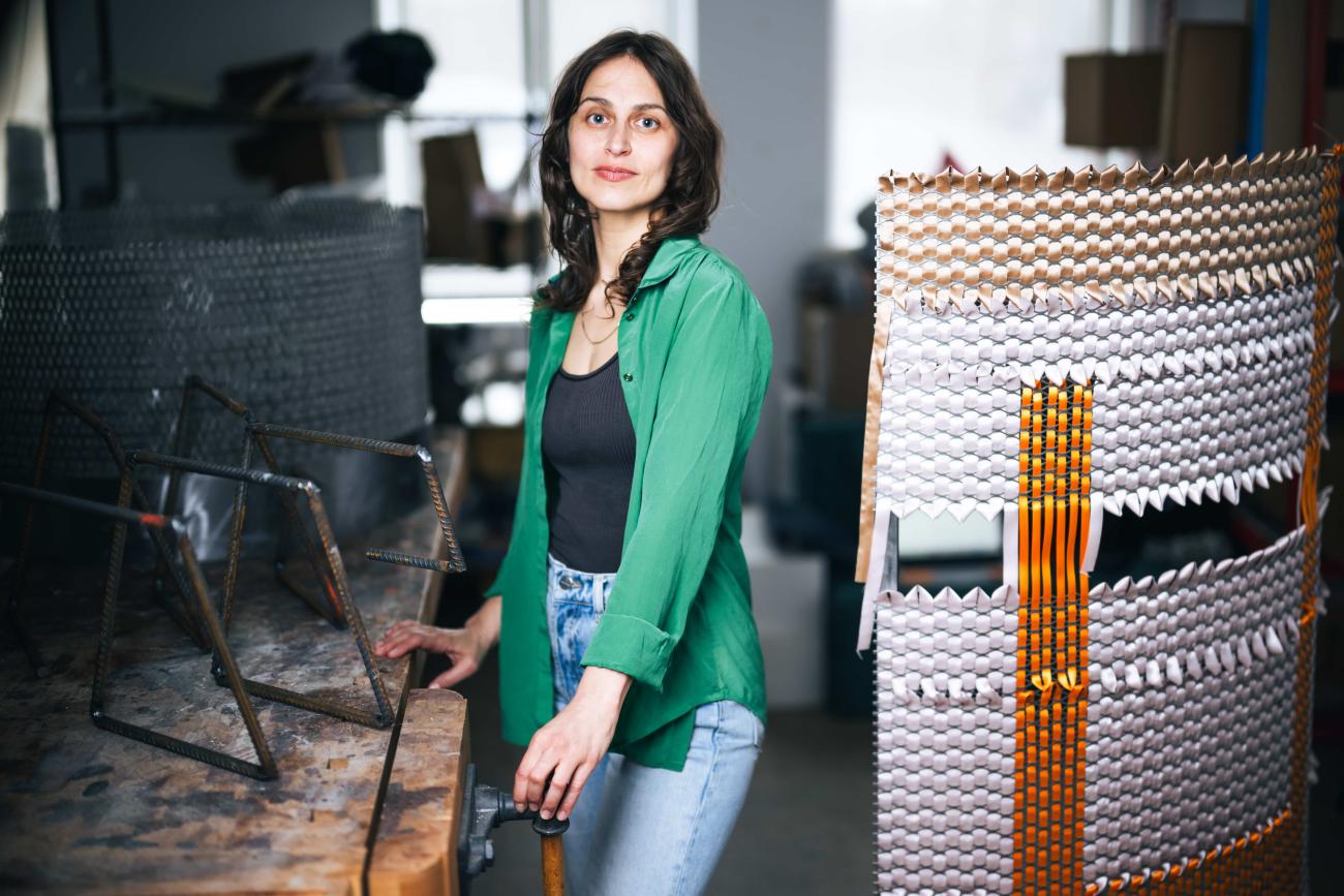 Alina Tenser stands between a rustic wooden workbench holding metal rebar structures and a large, curved sculpture featuring woven metallic and orange ribbons.